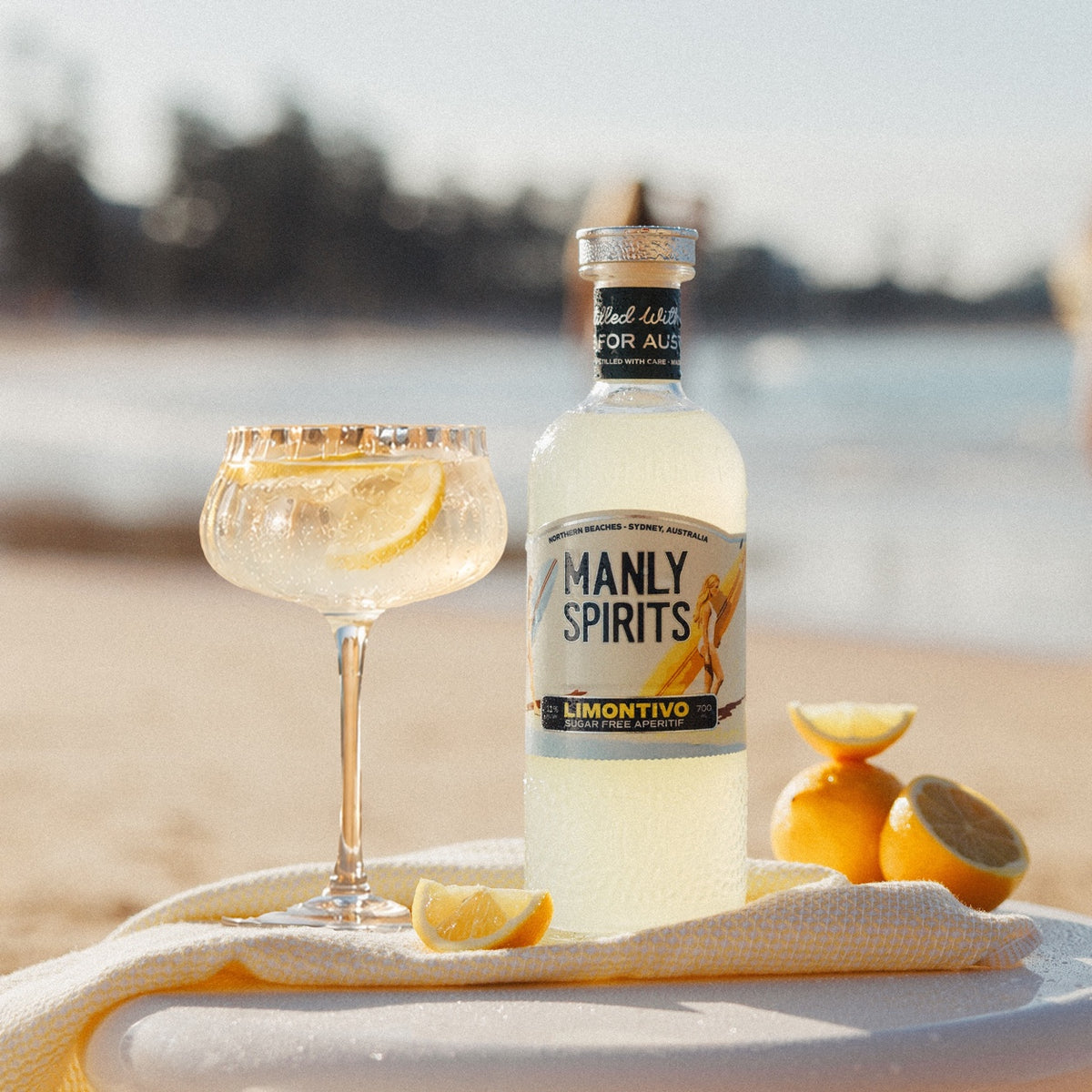Manly Spirits Limontivo Sugar Free Aperitivo on a towel at the beach with a coupe of Limontivo Spritz and cut lemons, Manly Beach behind