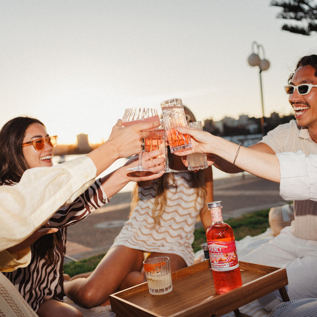 Friends toasting at a harbourside picnic at sunset, with a bottle of Lychee & Dragonfruit Liqueur on the tray