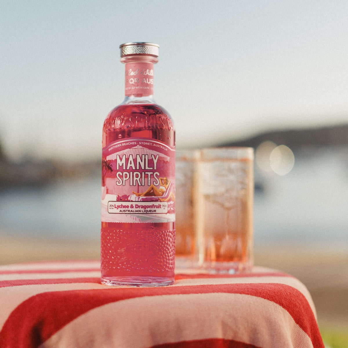 Manly Spirits Lychee & Dragonfruit Liqueur on a red and pink striped towel with two pink highballs, harbour behind