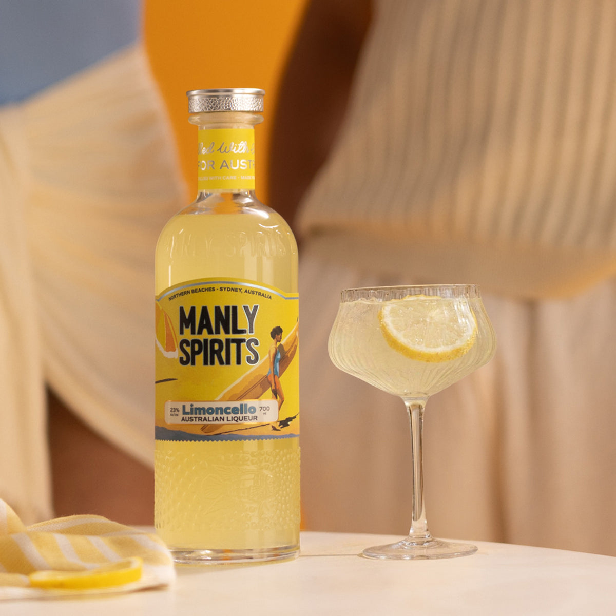 Manly Spirits Zesty Limoncello bottle beside a coupe spritz with a lemon wheel on a white table, yellow striped towel and soft orange and cream background.