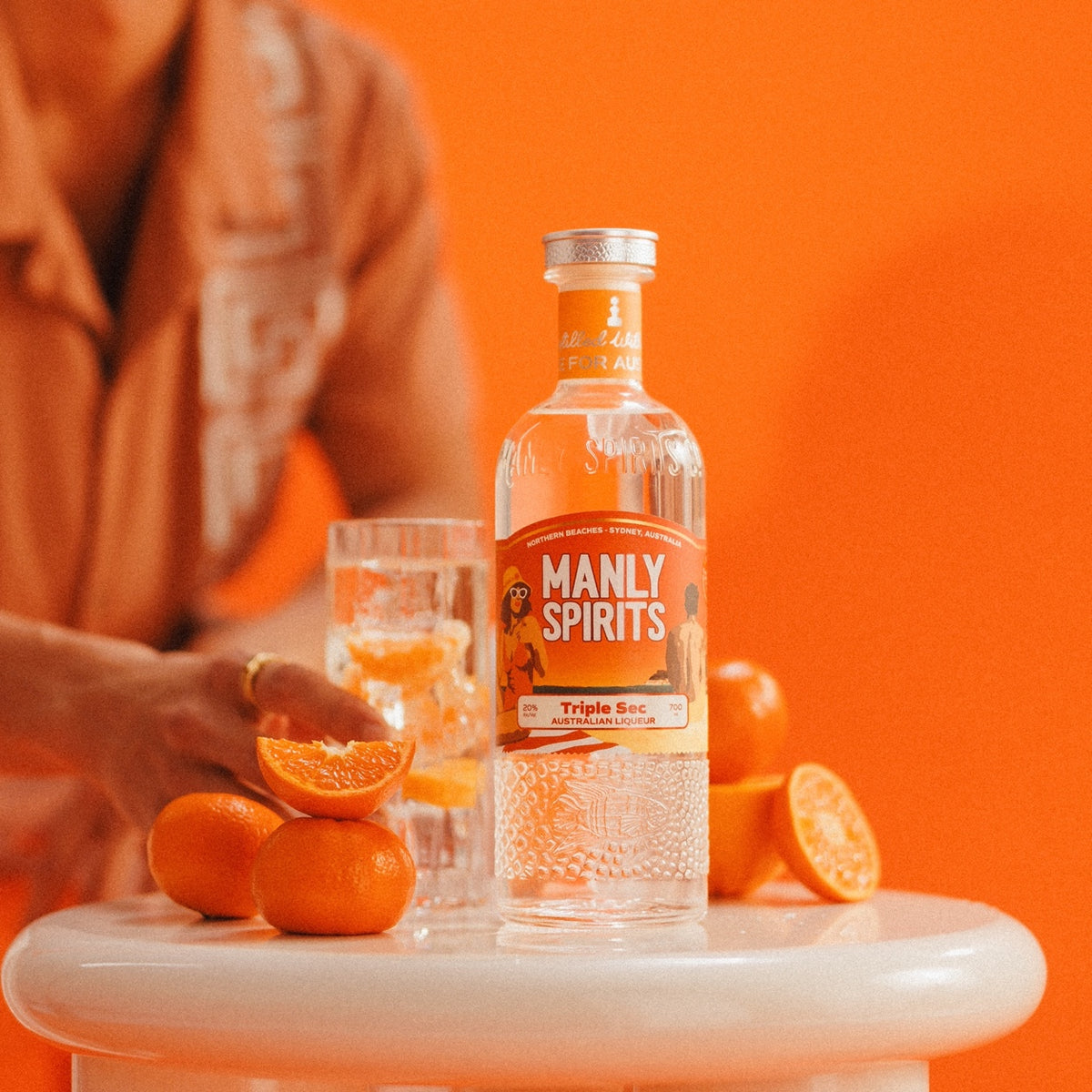 Manly Spirits Mandarin Triple Sec on a white side table with fresh mandarins, a man's hand resting on a mandarin half beside the highball, orange wall behind