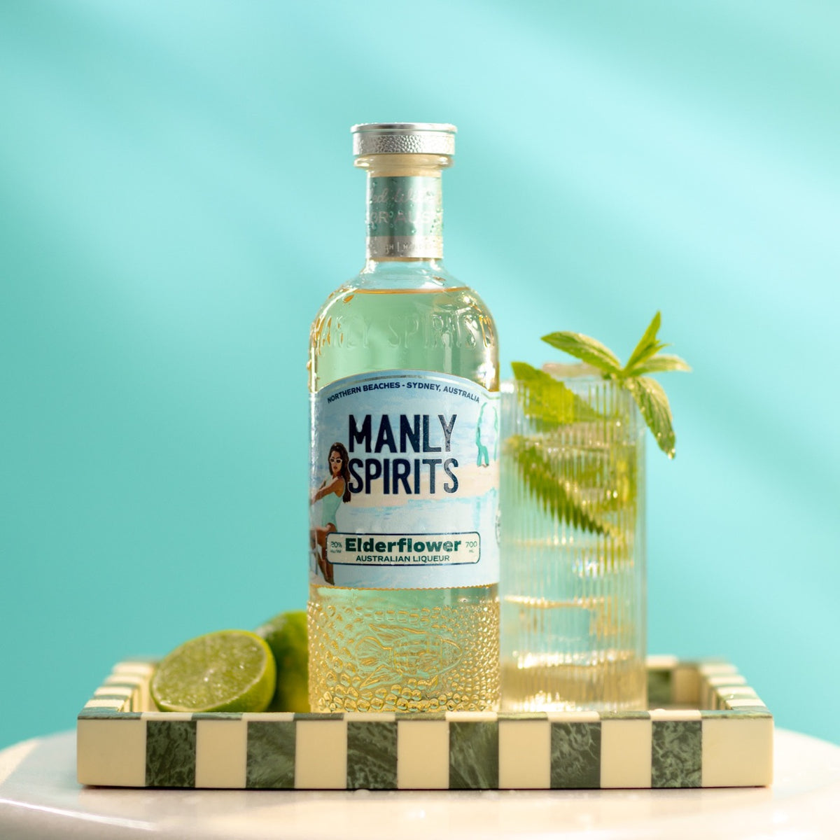 Manly Spirits Elderflower Liqueur bottle on a green and cream checkered tray with lime halves and a ribbed glass of Hugo Spritz with mint, turquoise wall behind