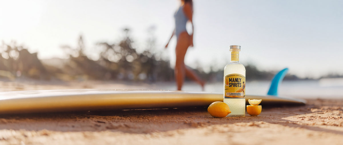 A bottle of Manly Spirits Zesty Limoncello on the sand beside fresh lemons and a surfboard, at the beach on a sunny day.