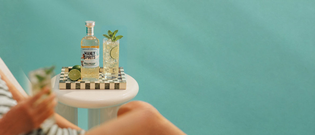 Elderflower Liqueur, limes and a Hugo Spritz in a tall glass with mint and lime on a checkered tray against a turquoise wall