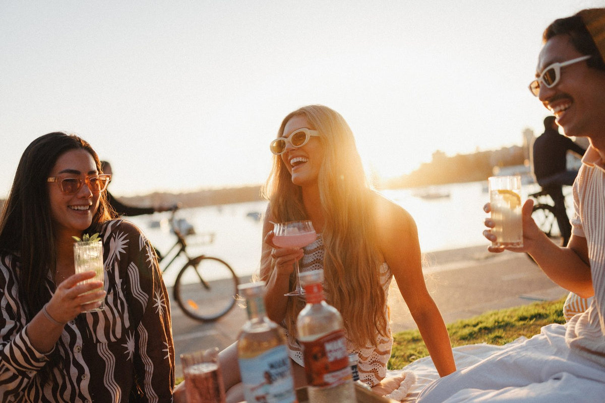 Three friends laughing at a harbourside picnic at sunset, one holding a Hugo Spritz in a tall ribbed glass with mint