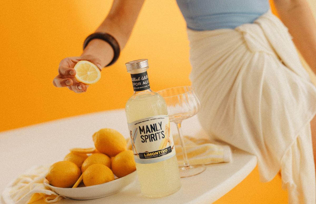 Squeezing a lemon beside a bottle of Limontivo and a bowl of lemons against a warm orange wall