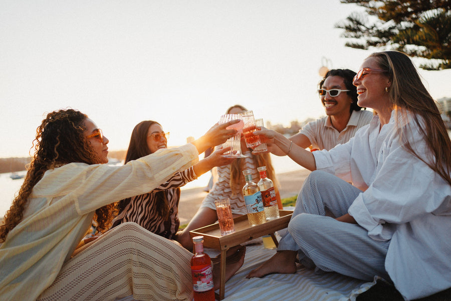 Friends toasting at sunset, with Lychee & Dragonfruit Liqueur in front and two more spritz liqueurs from the range on the tray