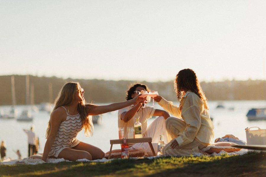 Three friends toasting with pink drinks on a picnic blanket by the harbour, with Lychee & Dragonfruit Liqueur beside the tray