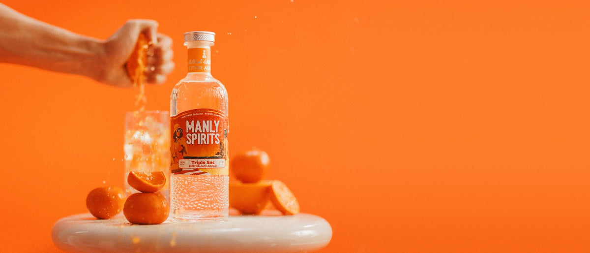 Mandarin Triple Sec beside fresh mandarins and a glass of ice as a hand squeezes a mandarin, against an orange wall