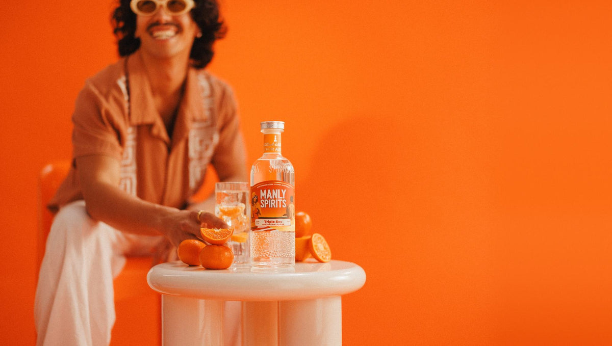 Mandarin Triple Sec, fresh mandarins and a tall glass over ice on a white side table against an orange wall