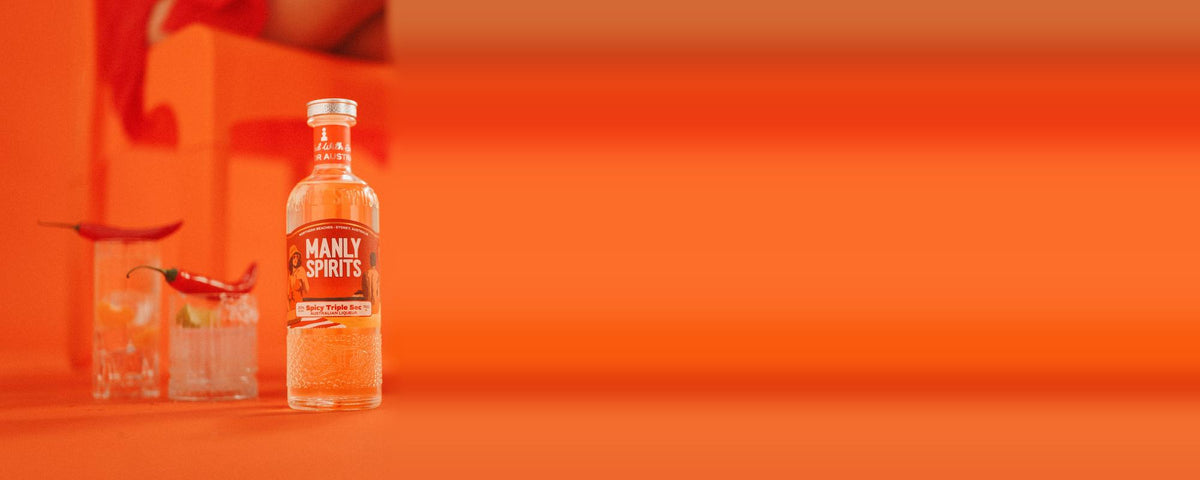 A bottle of Spicy Triple Sec on an orange set beside two iced drinks topped with fresh red chillies