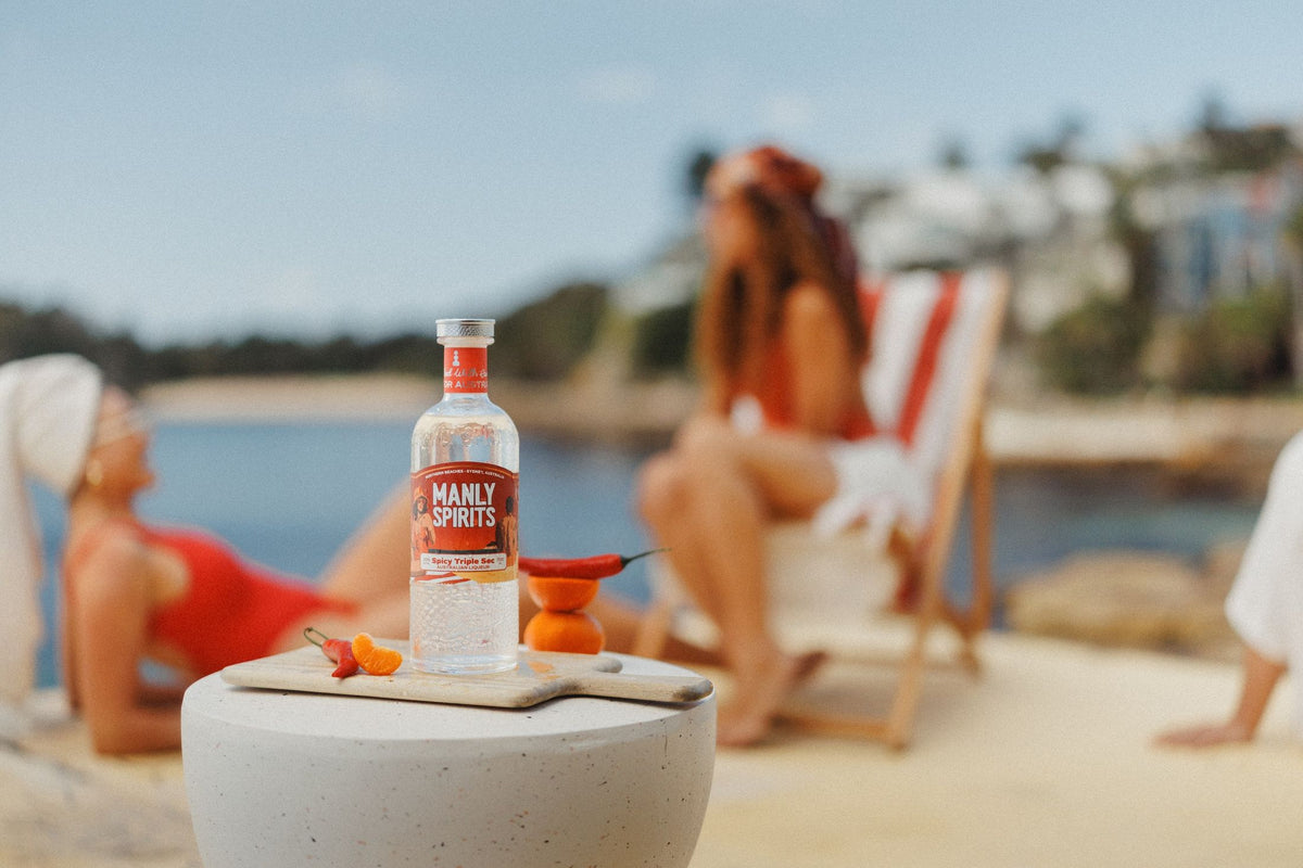 Spicy Triple Sec bottle with chillies and mandarins on a stone table by the harbour, friends relaxing behind