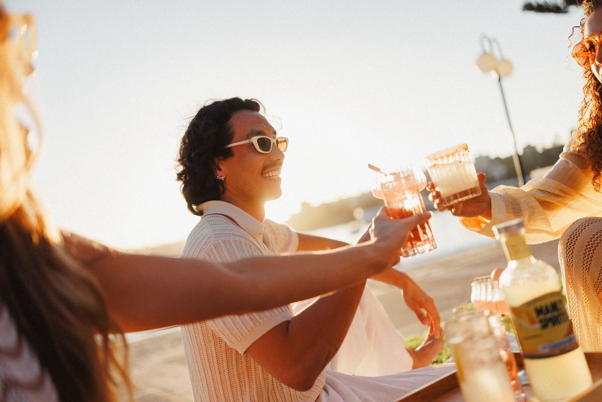 Friends raising their glasses at a waterside table at sunset, with a bottle of Zesty Limoncello in the foreground