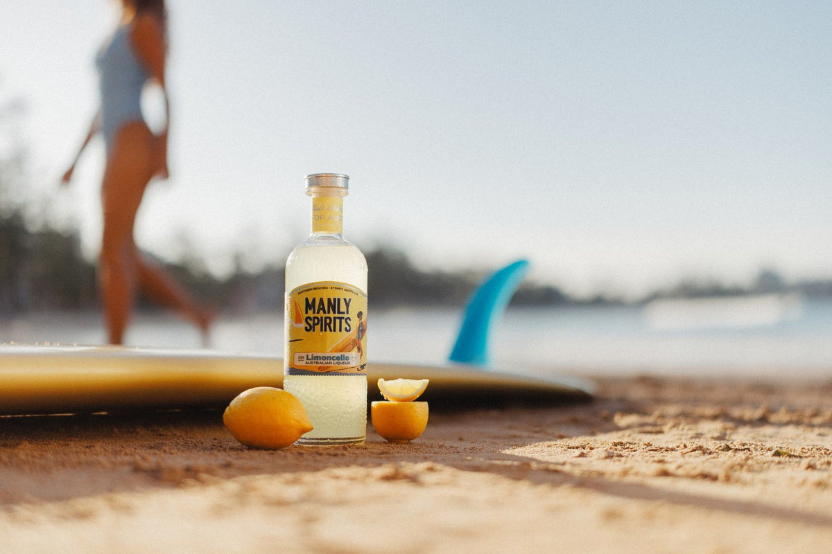 A bottle of Zesty Limoncello standing in the sand beside fresh lemons, with a surfboard and the beach behind
